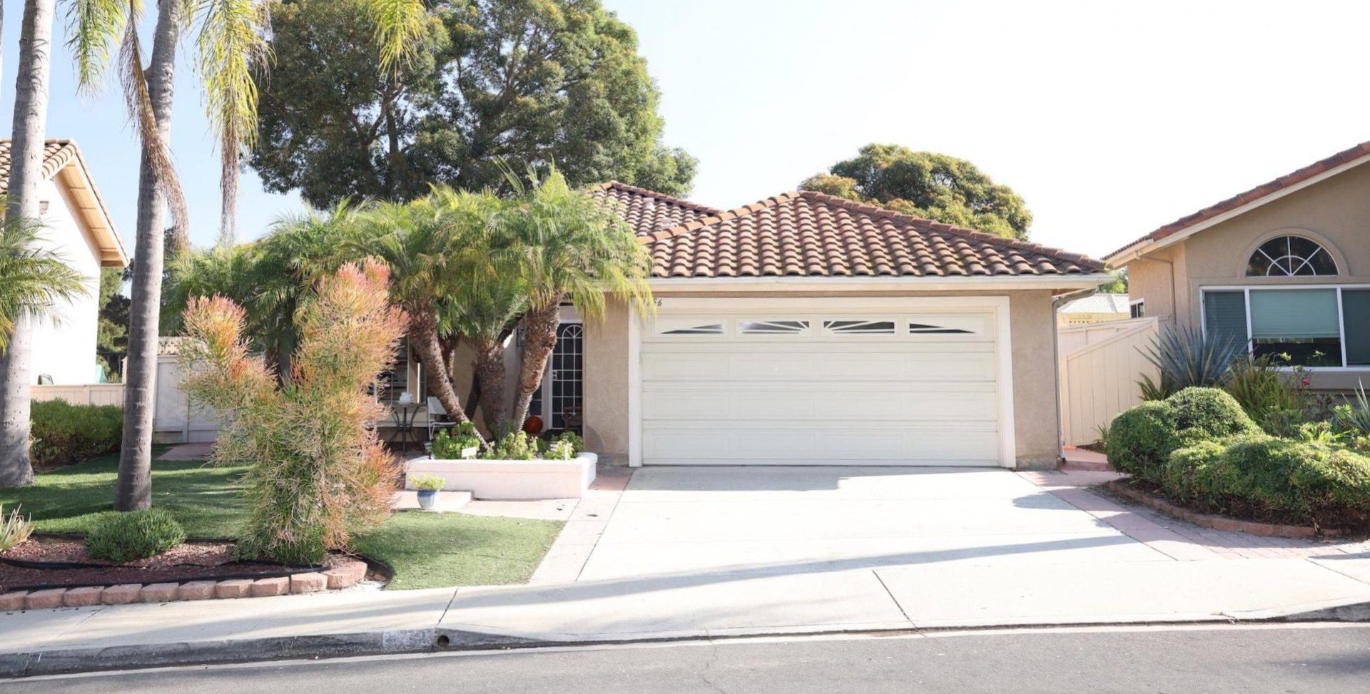 Modern single-story suburban house with white garage door, tiled roof, and landscaped front yard with palm trees and shrubs